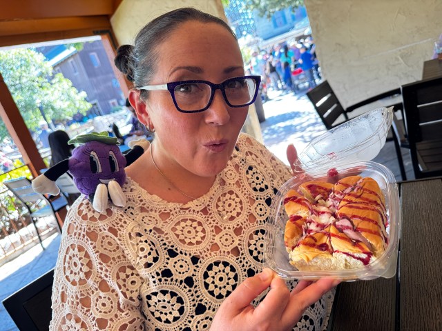 A woman with glasses holds a dessert in a takeout container, making a playful expression. She has a purple plush toy on her shoulder, and the setting features outdoor dining at a festival.