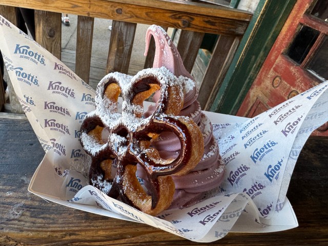 A dessert featuring a soft swirl of boysenberry ice cream topped with a large chocolate-dipped bunuelo, presented in a tray with Knott's Berry Farm branding.