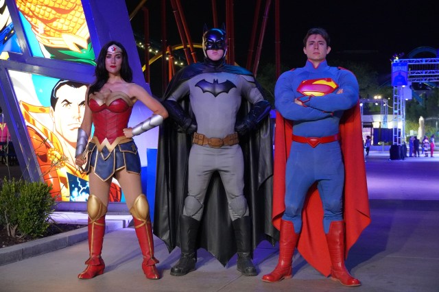 Three costumed characters stand in a brightly lit area: Wonder Woman, Batman, and Superman, with vibrant superhero-themed backdrops.